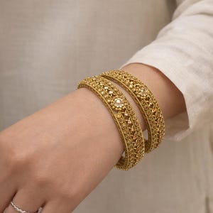 Puede incluir: Dos pulseras doradas con intrincados diseños y acentos brillantes se lucen en una muñeca. Las pulseras tienen un diseño detallado con pequeñas piedras incrustadas. La muñeca también tiene un anillo plateado.