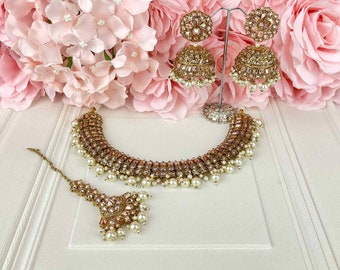 Pode incluir: Um conjunto de joias de noiva indianas em ouro e branco com um colar, brincos e um maang tikka. O colar é adornado com pérolas e pedras brilhantes. Os brincos são de estilo pendente com forma redonda e o maang tikka é um adorno tradicional para a testa.