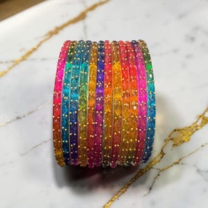 Könnte beinhalten: Ein Stapel bunter Armreifen in Regenbogenfarben, von Rot bis Violett. Jeder Armreif ist mit kleinen, goldfarbenen Punkten verziert. Die Armreifen liegen auf einer weißen Oberfläche mit goldenen Adern.