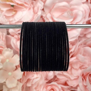 May include: A stack of black velvet bangles, approximately 18 in total, are displayed against a backdrop of pink fabric flowers.