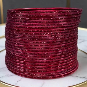 Op de afbeelding: Een stapel rode armbanden met glitteraccenten. De armbanden zijn gestapeld op een wit en goudkleurig oppervlak. De armbanden zijn dieprood van kleur en lijken van metaal te zijn.