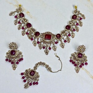 May include: A complete jewelry set featuring a necklace, earrings, and a headpiece. The set is gold-toned with burgundy-colored stones and clear crystals. The necklace and earrings have a floral design with dangling burgundy beads. The headpiece has a central burgundy stone.