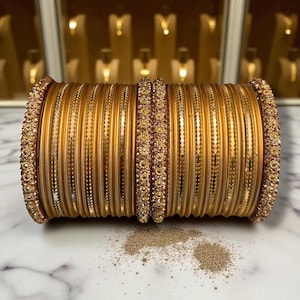 Puede incluir: Un conjunto de pulseras doradas, con varios diseños. Algunas pulseras están adornadas con pequeños adornos brillantes, mientras que otras tienen un acabado liso y pulido. Las pulseras están apiladas, mostrando sus intrincados detalles y su tono dorado.