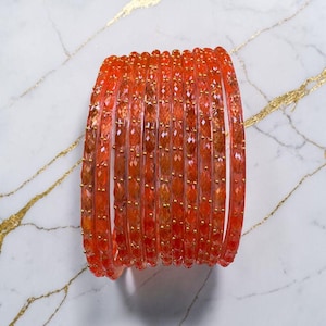 Op de afbeelding: Een stapel oranje armbanden met een facetontwerp. De armbanden hebben een levendige oranje kleur met kleine goudkleurige accenten. De sieraden worden gepresenteerd op een wit marmeren oppervlak met gouden aderen.