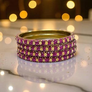 Op de afbeelding: Vier gouden armbanden, elk versierd met magenta en gele edelstenen. De armbanden zijn op elkaar gestapeld, wat het ingewikkelde ontwerp van de edelstenen laat zien. De edelstenen zijn gelijkmatig verdeeld over de omtrek van elke armband.