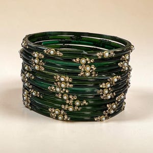 May include: A stack of dark green translucent bangles featuring gold-toned metallic embellishments and small white faux pearls arranged in floral patterns along the outer surface of each individual circular bracelet.
