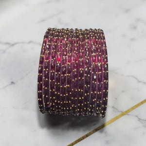 以下が含まれることがあります： 金色のアクセントが付いた紫色のバングルのスタック。バングルは深い紫の色で、ファセットデザインで、小さな金色のビーズが散りばめられています。背景は、金色の線が入った白い大理石の表面です。