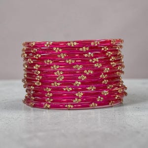 May include: A stack of twelve translucent fuchsia pink bangles, each featuring a repeating pattern of small, gold-toned floral embellishments set with tiny clear crystals arranged in a horizontal line around the circumference of the bracelets.