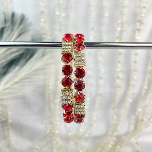 May include: Two gold-tone bracelets with red and clear rhinestones. The bracelets are linked together and hanging from a silver rod.