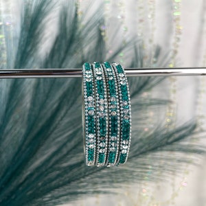 Peut inclure: Un ensemble de cinq bracelets verts et argentés. Chaque bracelet est orné de rangées alternées de pierres vert émeraude et transparentes scintillantes. Les bracelets sont présentés sur une barre argentée sur un fond flou.