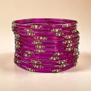 Op de afbeelding: Een stapel doorschijnende magenta armbanden, elk voorzien van kleine, gelijkmatig verdeelde goudkleurige metalen accenten ingelegd met kleine witte kunstparels. De ronde armbanden zijn gerangschikt in een verticale kolom.