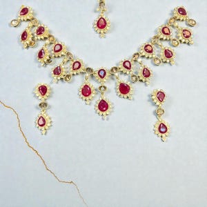 May include: A gold-toned jewellery set featuring a necklace, a pair of drop earrings, and a maang tikka. Each piece is adorned with teardrop-shaped red stones surrounded by small clear crystals, arranged in a repeating floral-inspired pattern.