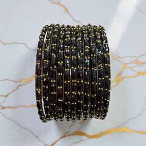 May include: A stack of twelve black bangles featuring a faceted, textured surface. Each bangle is accented with small, evenly spaced gold-coloured beads embedded along the outer edge, creating a uniform and repeating pattern across the entire set.