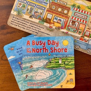 May include: A blue and yellow children's book titled "A Busy Day on the North Shore". The cover features a colorful illustration of a beach scene with waves, a small island, and a lighthouse. The text "Written by Jim Mayfield Illustrated by Monika Iwata" is at the bottom of the cover.