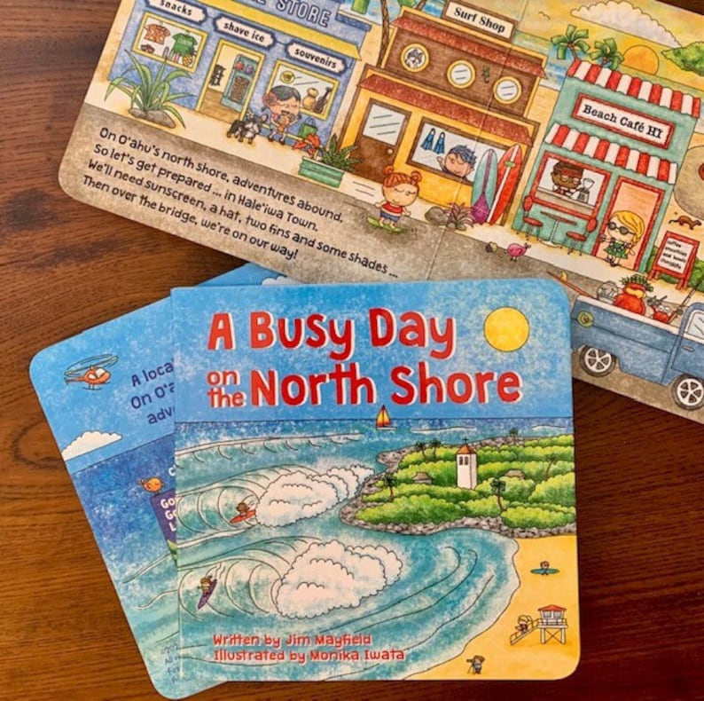 May include: A blue and yellow children's book titled "A Busy Day on the North Shore". The cover features a colorful illustration of a beach scene with waves, a small island, and a lighthouse. The text "Written by Jim Mayfield Illustrated by Monika Iwata" is at the bottom of the cover.