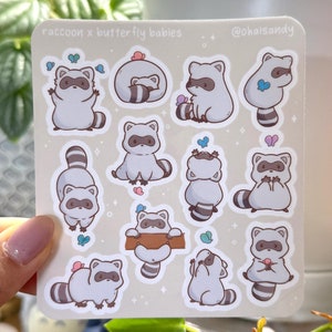 May include: A sticker sheet featuring cartoon raccoons with butterfly accents. The text "raccoon x butterfly babies" is printed at the top. The raccoons are in various poses, with hearts and butterflies. The background is a light beige color.