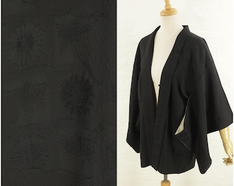 May include: A black haori jacket featuring a subtle, tonal woven pattern of floral and geometric shapes. The garment has wide, loose sleeves, an open front design, and a light-colored interior lining visible at the edges of the front panels.