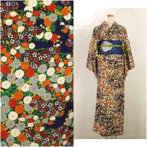 以下が含まれることがあります： オレンジ、白、グレーの花柄が紺色の背景に施された日本の着物。マネキンに着物が展示されており、青い帯と装飾的なアクセントが付いています。