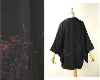 May include: Black kimono-style jacket with a floral design. The jacket features a loose fit with wide sleeves and a collar. The floral pattern is in shades of red and is subtly textured against the black fabric. The jacket is displayed on a mannequin.
