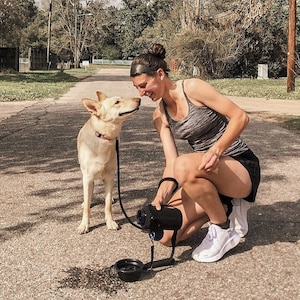 May include: A woman kneels on a paved road, interacting with a light-coloured dog on a lead. The dog wears a red collar. A black water bowl and a black object are on the ground. The background includes trees, houses, and a cloudy sky.
