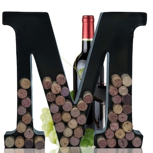 May include: Black metal letter M wine bottle holder with cork stoppers. The letter is filled with cork stoppers of various sizes and colors. A bottle of red wine is in the background.