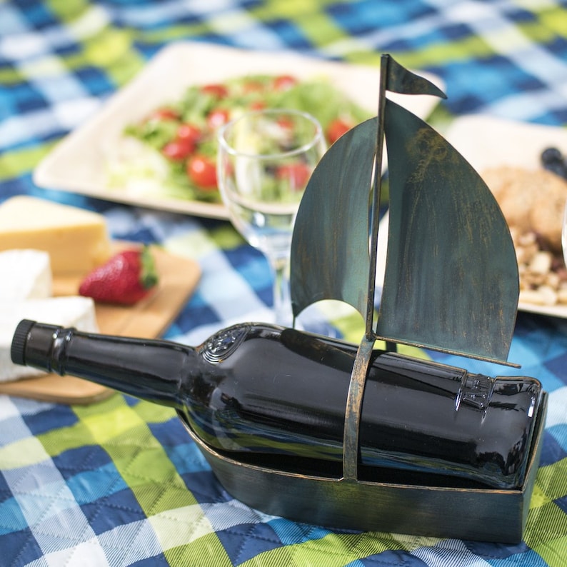 Puede incluir: Un porta botellas de vino de metal con forma de velero con un acabado azul oscuro y verde. El porta botellas sostiene una botella de vino de vidrio oscuro.