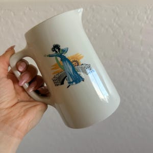 May include: White ceramic pitcher with a handle, featuring a vintage illustration of a woman in a blue dress, a building, and a sun. The pitcher has a curved spout and a large handle.