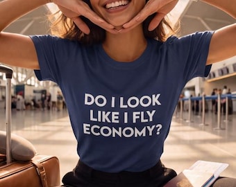 May include: A navy blue t-shirt with the white text DO I LOOK LIKE I FLY ECONOMY? worn with black trousers and white sneakers. A brown suitcase with a gray neck pillow and a passport with boarding passes are positioned nearby in an airport.