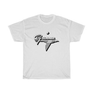 May include: White t-shirt featuring a black graphic design with the word "Grumman" in a stylized font, accompanied by an image of a bird in flight. The shirt has a crew neck and short sleeves.