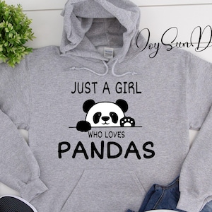 May include: A gray hooded sweatshirt with the text "JUST A GIRL WHO LOVES PANDAS" and a panda graphic. The sweatshirt is paired with blue jeans, black sneakers, and a white mug. A small green plant is also visible.
