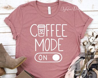 以下が含まれることがあります： 白い文字で「COFFEE MODE ON」と書かれた、薄紫色のTシャツ。ハートの付いたスタイリッシュなコーヒーカップのアイコンが描かれている。Tシャツは白い木製の表面に置かれ、茶色のスカーフと茶色のブーツが置かれている。