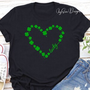 May include: Black t-shirt with a green shamrock heart design and the word "lucky" in cursive. The shirt is styled with denim shorts and white trainers. The image has a St. Patrick's Day theme.