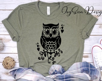 May include: A green t-shirt with a black silhouette of an owl perched on a branch with hearts and leaves. The text "Joy Sun Designs" is printed in the top right corner of the shirt.