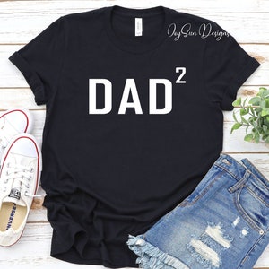 May include: Black t-shirt with white text that reads "DAD²". The shirt is laying on a white wooden surface with a pair of white sneakers and denim shorts.
