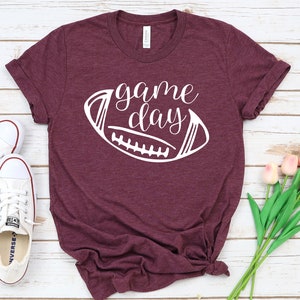 May include: A maroon t-shirt with a white graphic of a football and the text "game day" on it.