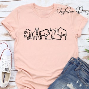 May include: A light pink t-shirt featuring a black line drawing of five bison. The shirt is paired with denim shorts and white canvas sneakers. The text "Joy Sun Design" is printed on the shirt.