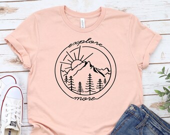 May include: A pink t-shirt with a black graphic of a mountain range, sun, and trees inside a circle with the text "explore more".