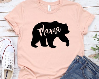 May include: A pink t-shirt featuring a black silhouette of a bear with the word "Mama" inscribed within the bear. The t-shirt is lying on a white wooden surface alongside a pair of white trainers and a pair of blue denim shorts.