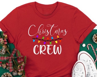 May include: A red t-shirt with the words "Christmas CREW" in white script and a string of colorful lights. White Converse sneakers and Christmas decorations are arranged around the shirt, suggesting a festive theme.