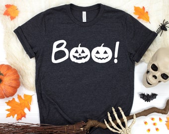 May include: Dark gray t-shirt with the word "Boo!" and two jack-o'-lanterns in white. The shirt is surrounded by Halloween decorations, including a pumpkin, leaves, a broom, and a skull.