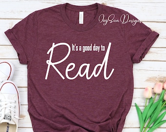 May include: A maroon short-sleeved t-shirt with white text that reads "It's a good day to Read" in a mix of sans-serif and script fonts. The shirt features a crew neckline and a heathered fabric texture, displayed flat with white sneakers and tulips.