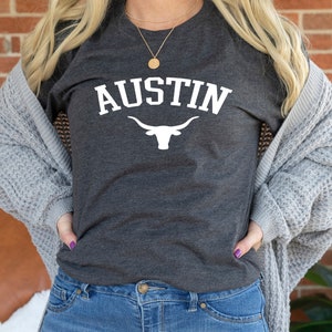 May include: A dark gray t-shirt with the word "AUSTIN" in white text and a white silhouette of a longhorn bull underneath. The person wearing the shirt is also wearing blue jeans and a gray cardigan.