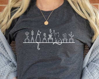以下が含まれることがあります： 暗いグレーのTシャツに、白い猫と鉢植えの植物のグラフィックがプリントされています。グラフィックは、鉢植えの植物の列と、それらと相互作用する猫を特徴としています。