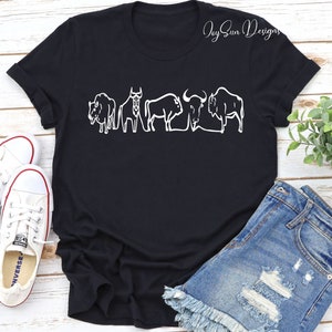 May include: Black t-shirt featuring a white outline design of five bison. The shirt has a crew neck and short sleeves. The text "Joy Sun Designs" is in the upper right corner. The shirt is paired with denim shorts and white sneakers.