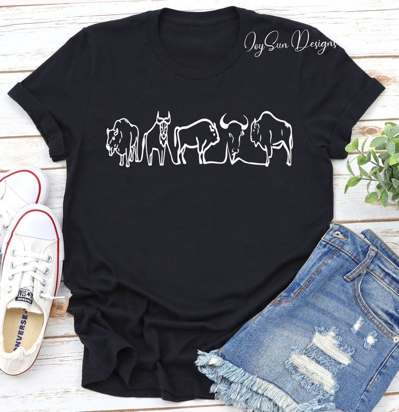 May include: Black t-shirt featuring a white outline design of five bison. The shirt has a crew neck and short sleeves. The text "Joy Sun Designs" is in the upper right corner. The shirt is paired with denim shorts and white sneakers.