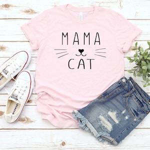 May include: Pink t-shirt with the text "Mama Cat" in black lettering. The text is accompanied by a stylized cat face with whiskers and a heart for a nose.