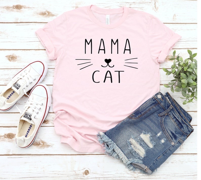 May include: Pink t-shirt with the text "Mama Cat" in black lettering. The text is accompanied by a stylized cat face with whiskers and a heart for a nose.