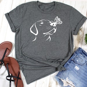May include: A dark gray t-shirt with a white silhouette of a dog's head sniffing a butterfly. The dog's head is facing the right side of the image.