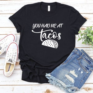 以下が含まれることがあります： 白い文字で「You had me at tacos」と書かれた黒いTシャツと、白い輪郭のタコの絵が描かれています。Tシャツは木製の表面に置かれ、白いスニーカーとデニムのショートパンツが置かれています。