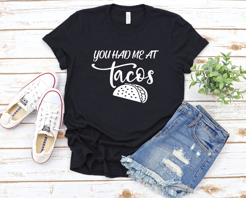 以下が含まれることがあります： 白い文字で「You had me at tacos」と書かれた黒いTシャツと、白い輪郭のタコの絵が描かれています。Tシャツは木製の表面に置かれ、白いスニーカーとデニムのショートパンツが置かれています。